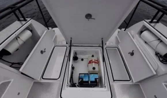 Open boat storage compartment featuring batteries, a fuel tank, and two side cabinets on a deck above water.
